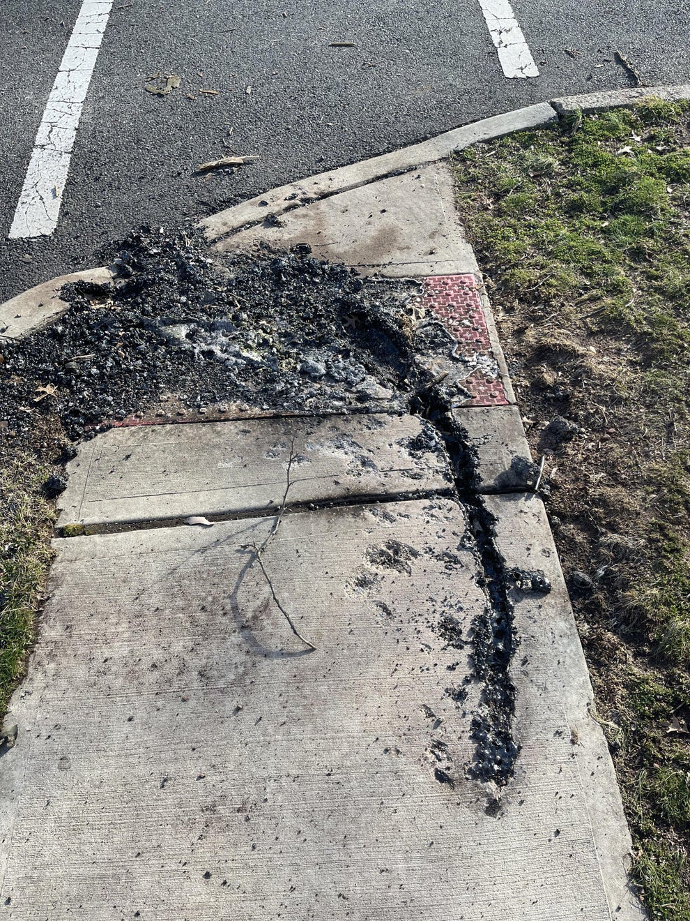 Damaged sidewalk and curb with scorched debris, possibly from a fire, surrounded by a road and grass area
