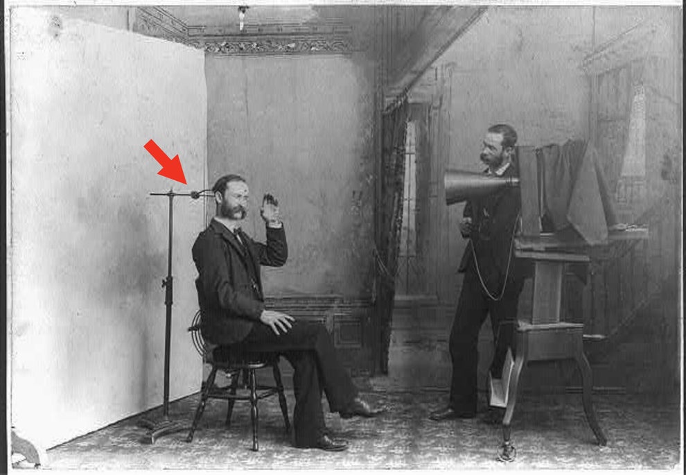 A man sits with earbuds beside an early phonograph, while a photographer captures the moment using a large vintage camera