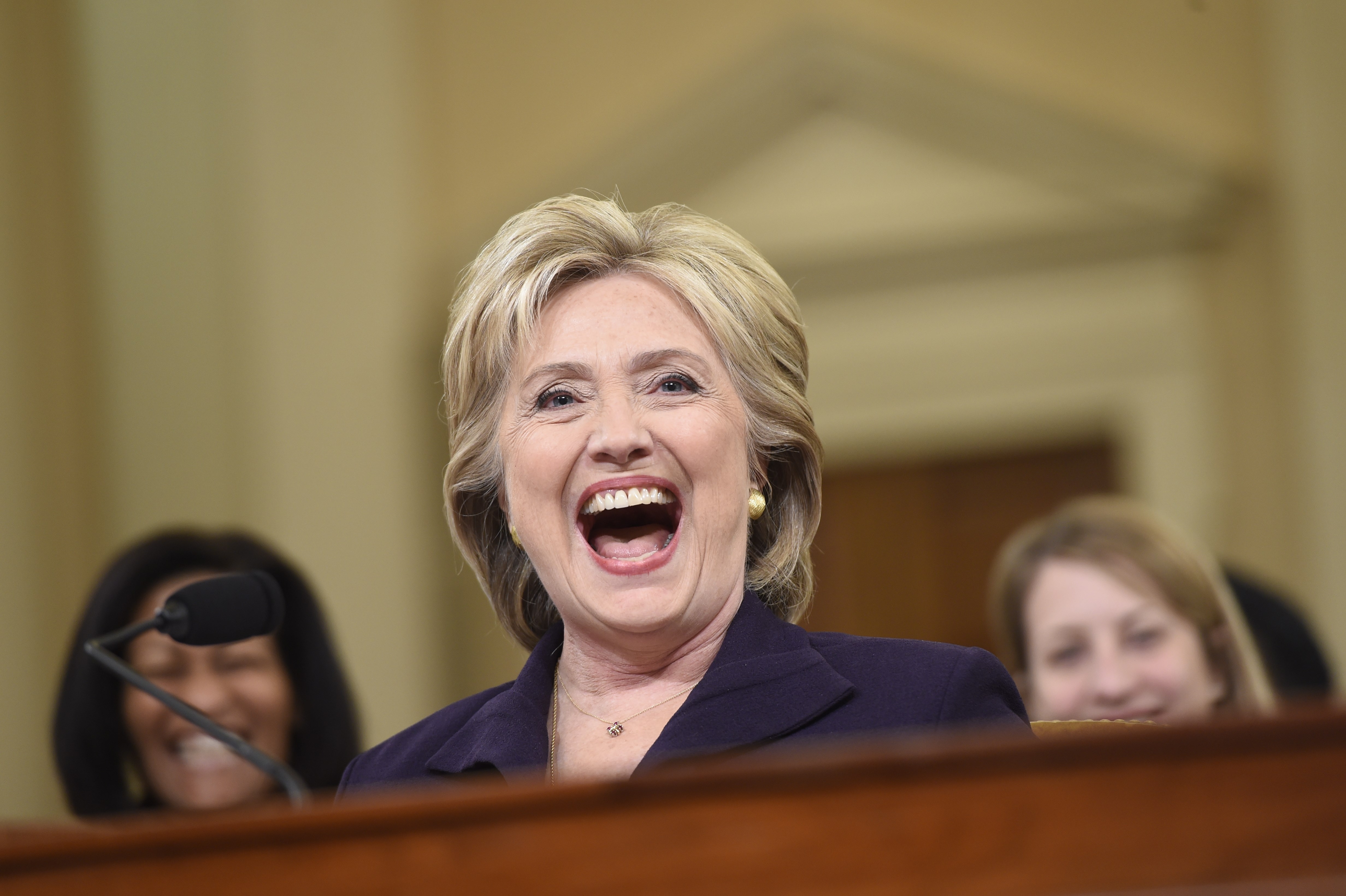 Smiling woman in a formal suit, speaking at an event, with others in the background