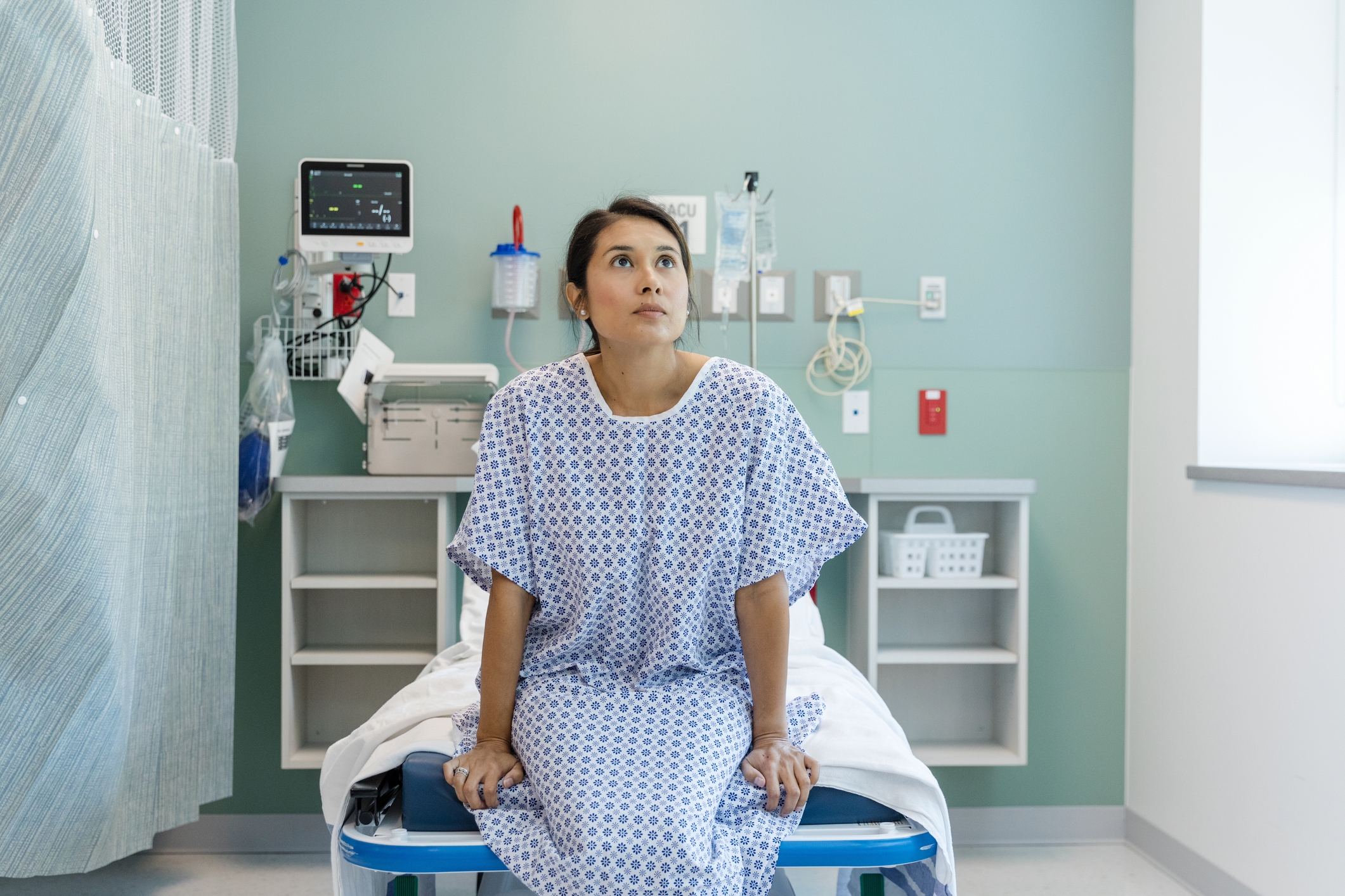 Person som sitter på en sykehusseng i en pasientkjole, ser oppover, i et medisinsk rom