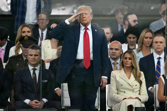 A man in a suit and red tie salutes while standing among seated attendees at a formal event
