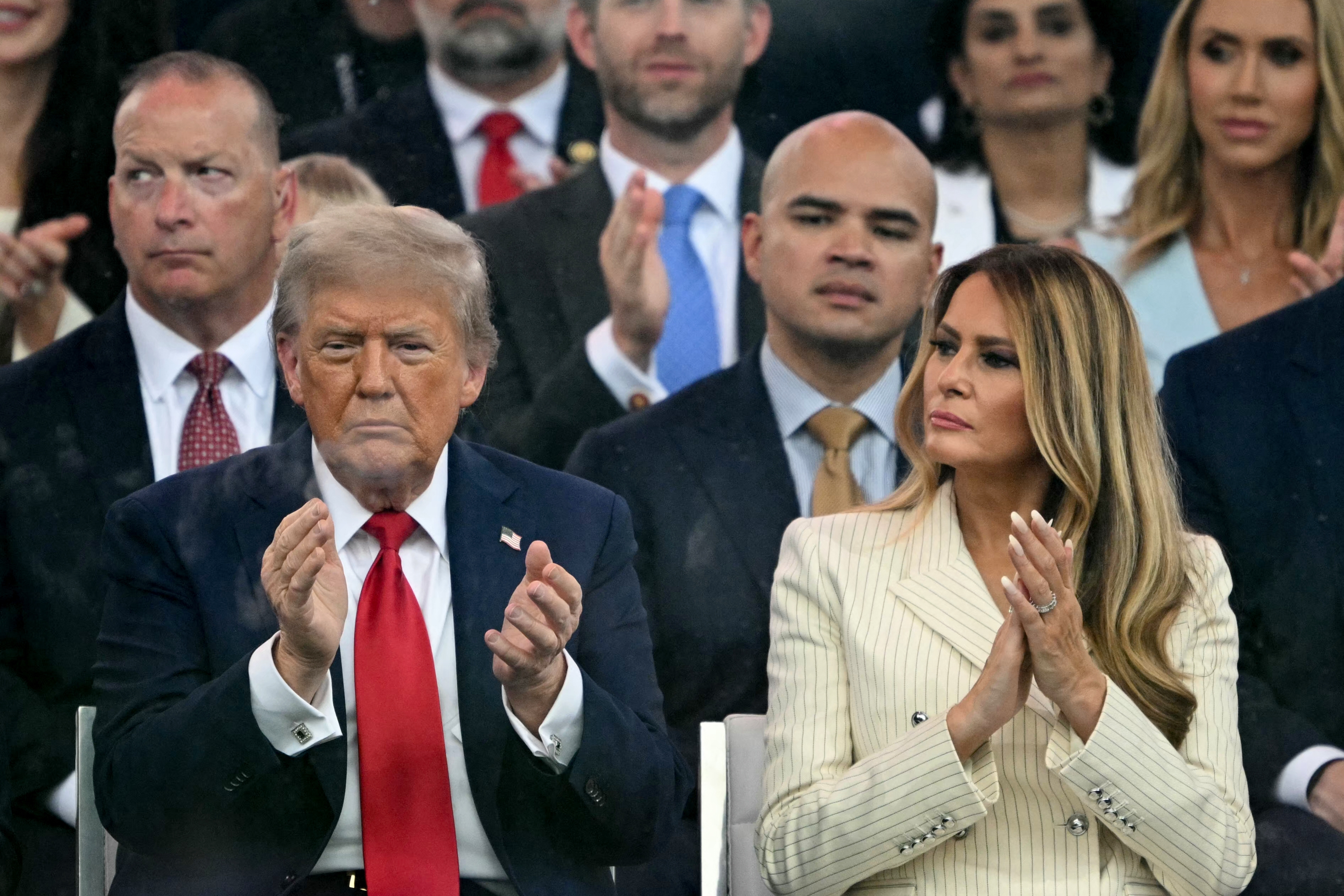 A man in a suit and red tie claps while sitting next to a woman in a pinstripe blazer at an event. Others are clapping in the background