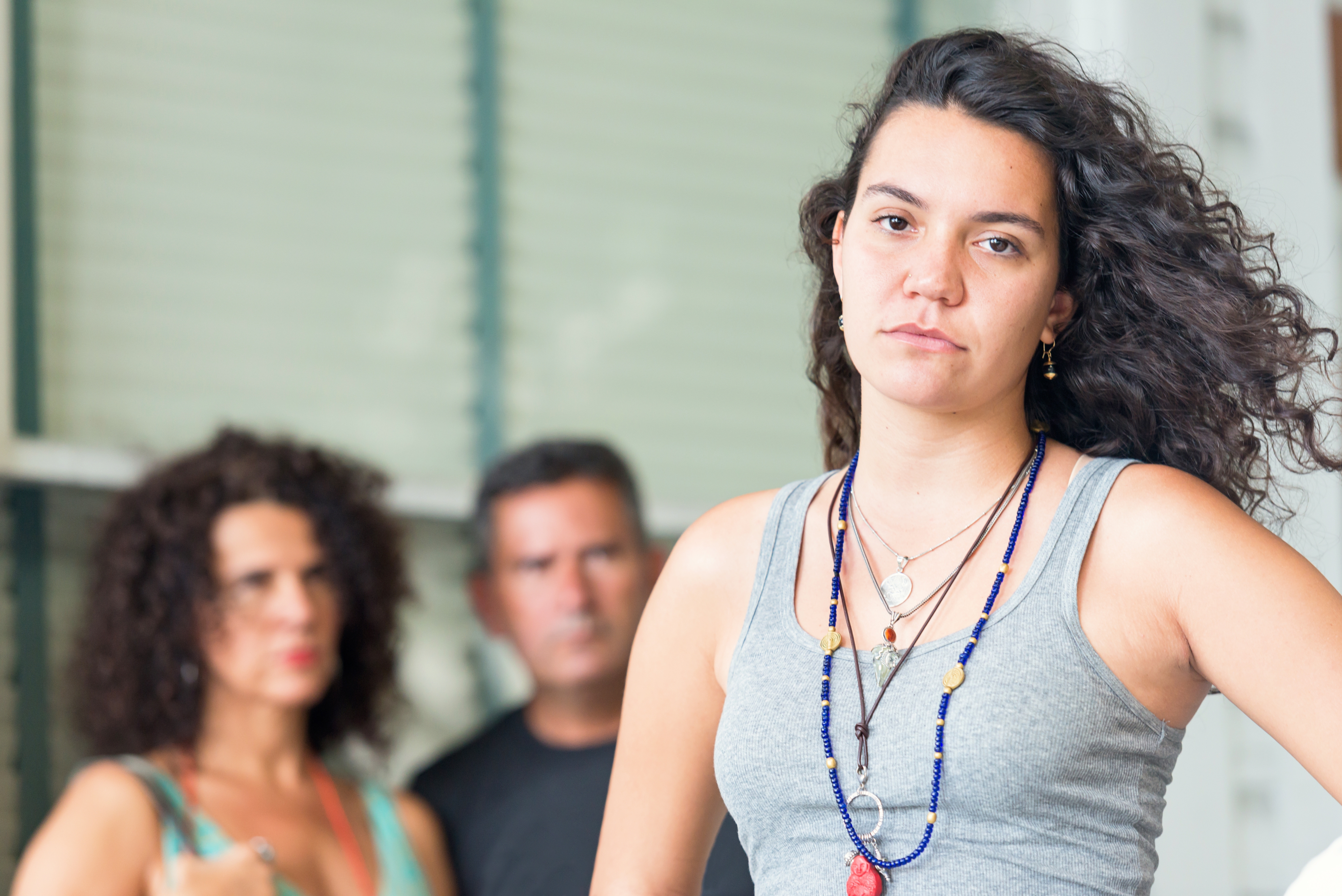 En kvinne i en tank top og lagdelte halskjeder ser trygt på kameraet, med to personer i den uskarpe bakgrunnen