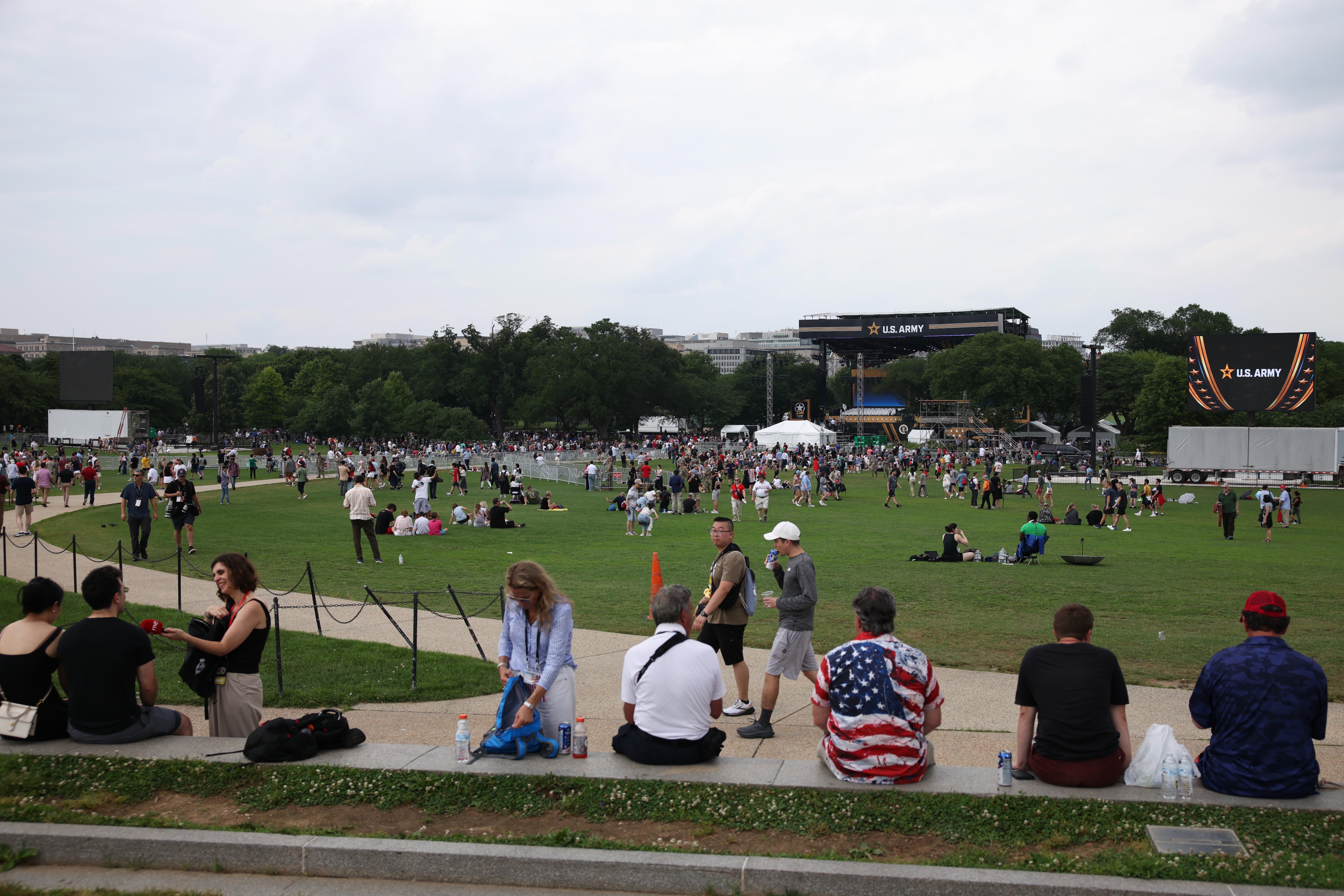 People gather on a large grassy area with scattered groups sitting and walking. A large screen displays &quot;U.S. Army&quot; in the background