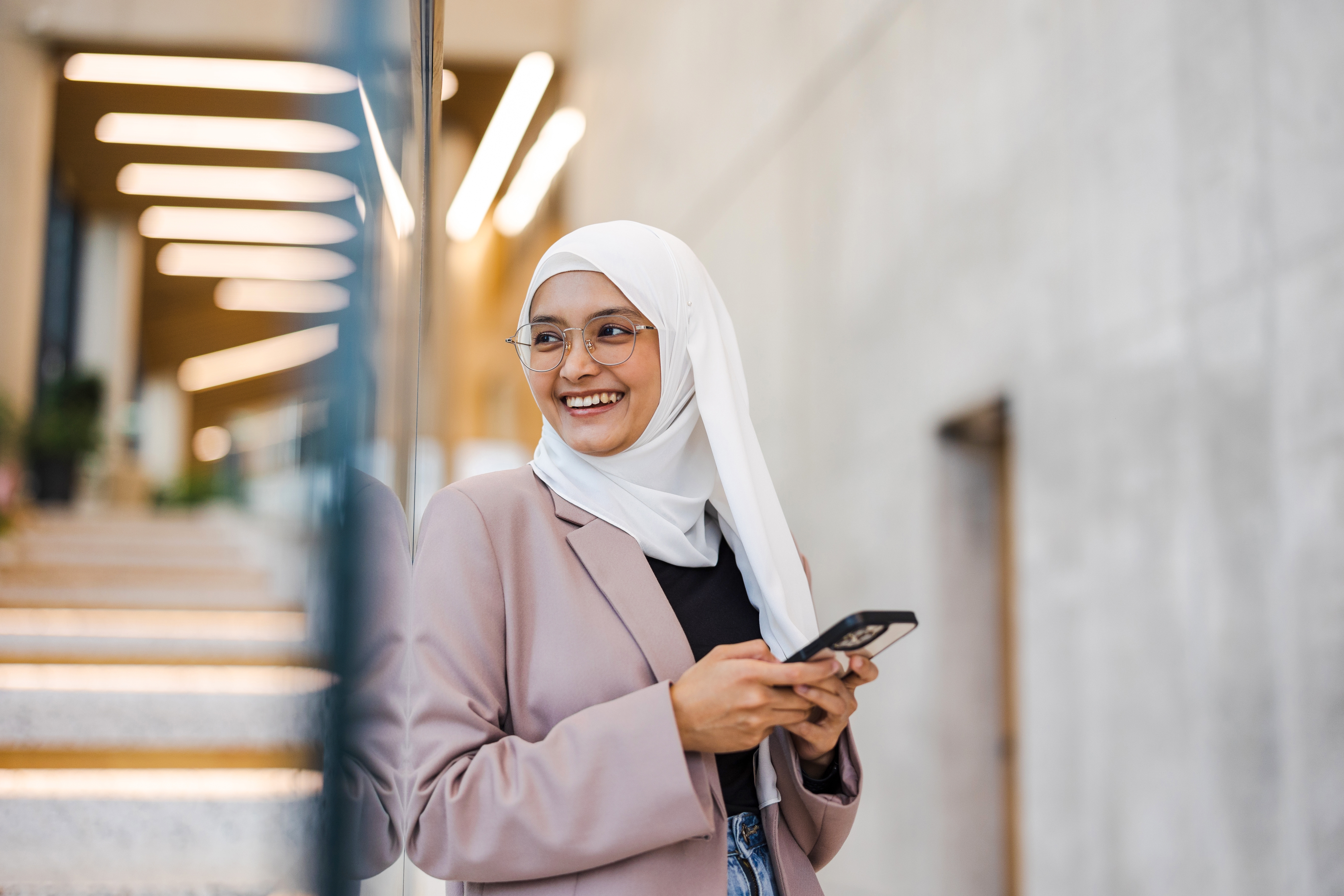 Person i en blazer og hijab smiler mens han holder en smarttelefon, og står innendørs i nærheten av et glass og betongvegg