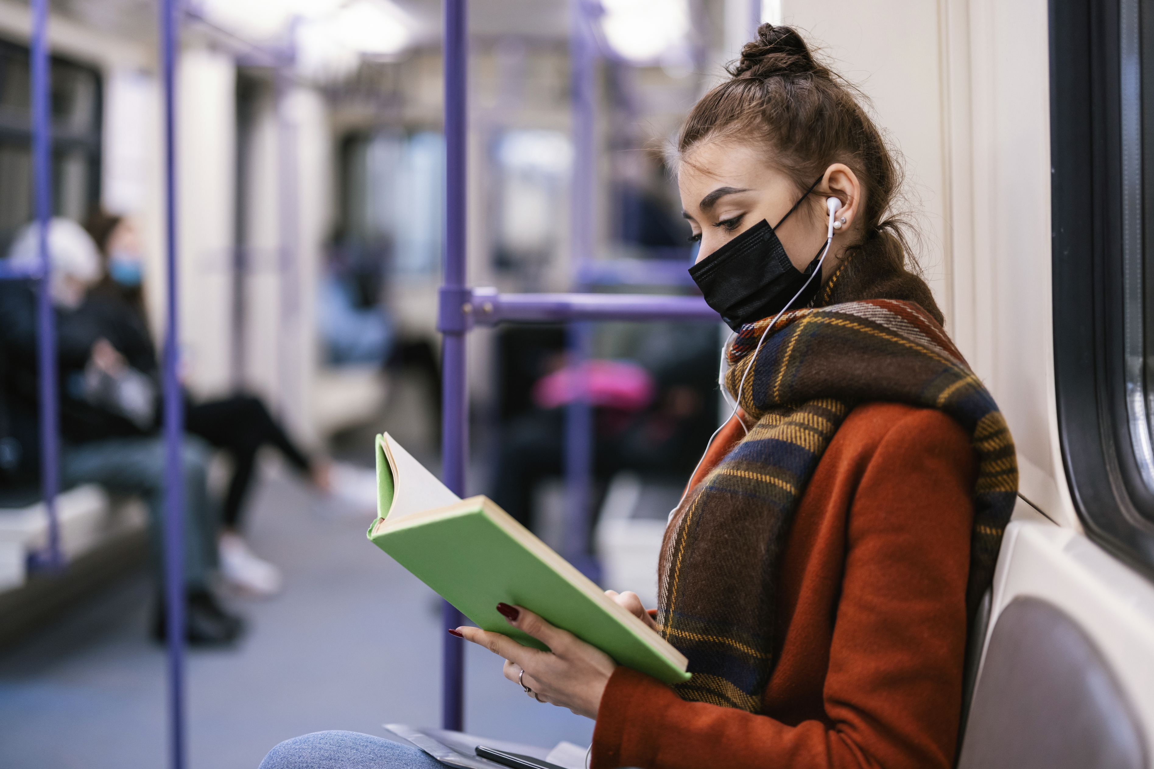 Person i en jakke og skjerf, iført en maske, sitter på offentlig transport og leser en bok og lytter til øretelefoner. Andre passasjerer er i bakgrunnen