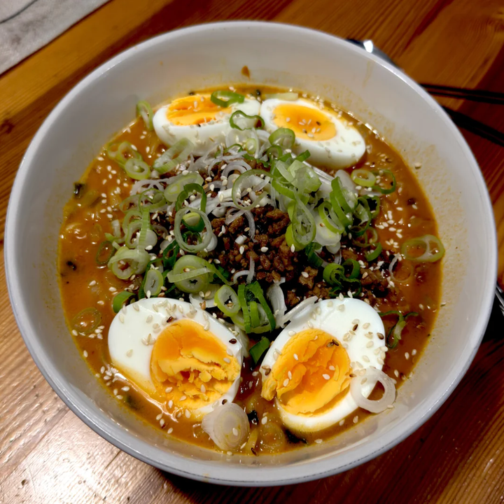 A bowl of Ramin with boiled egg slices, green onions, sesame seeds and ground meat on a wooden table