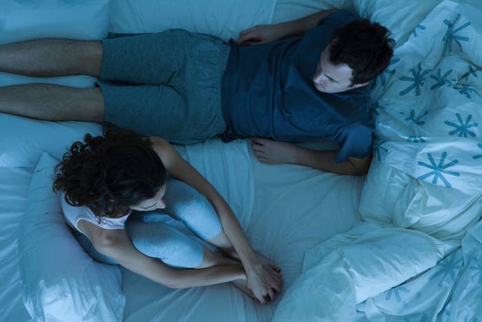 A couple in casual loungewear sits on a bed, appearing deep in conversation, suggesting intimacy and connection