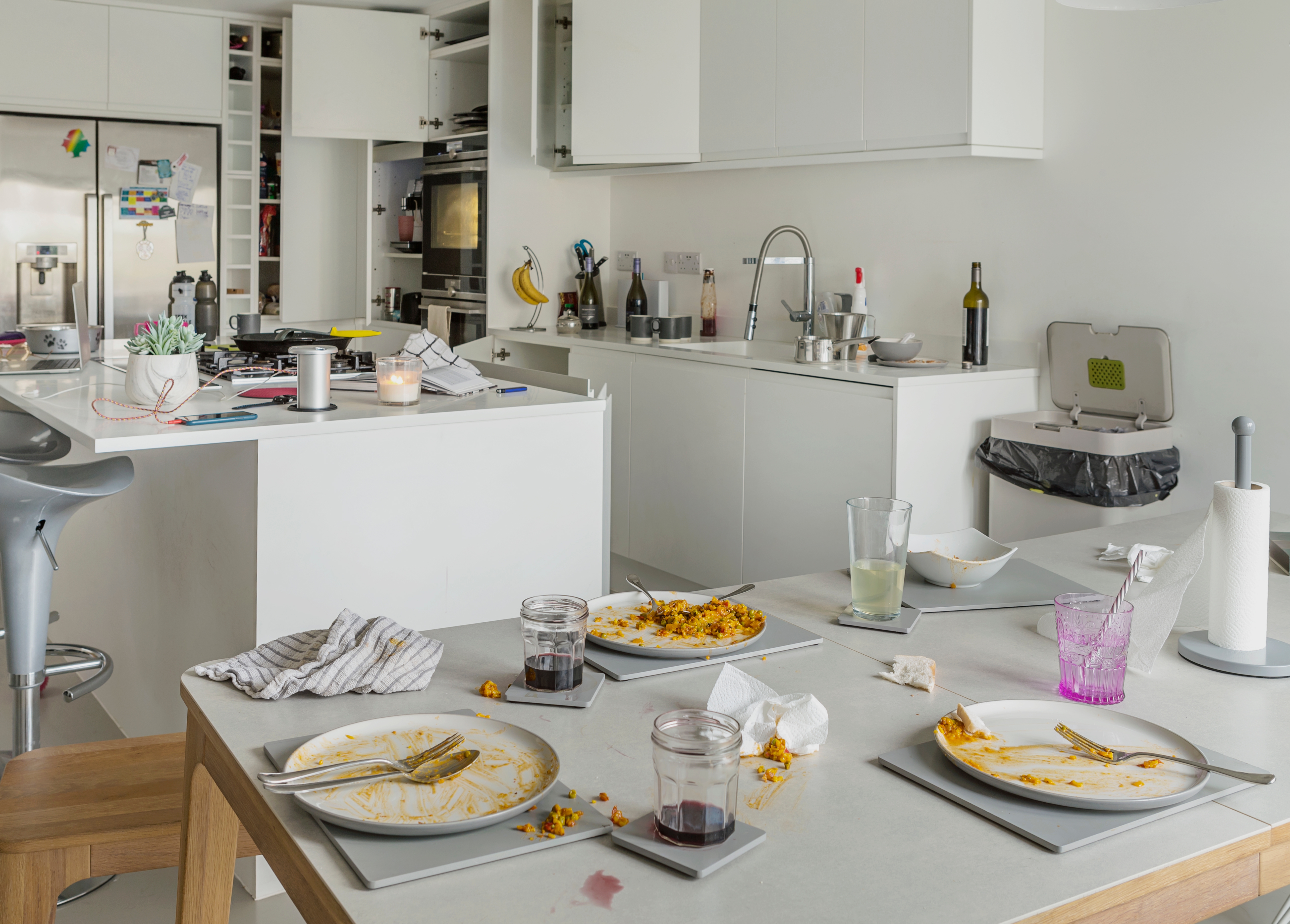 Messy kitchen with unwashed dishes and glasses on a table, leftover food on plates. The background shows a modern kitchen with various items scattered