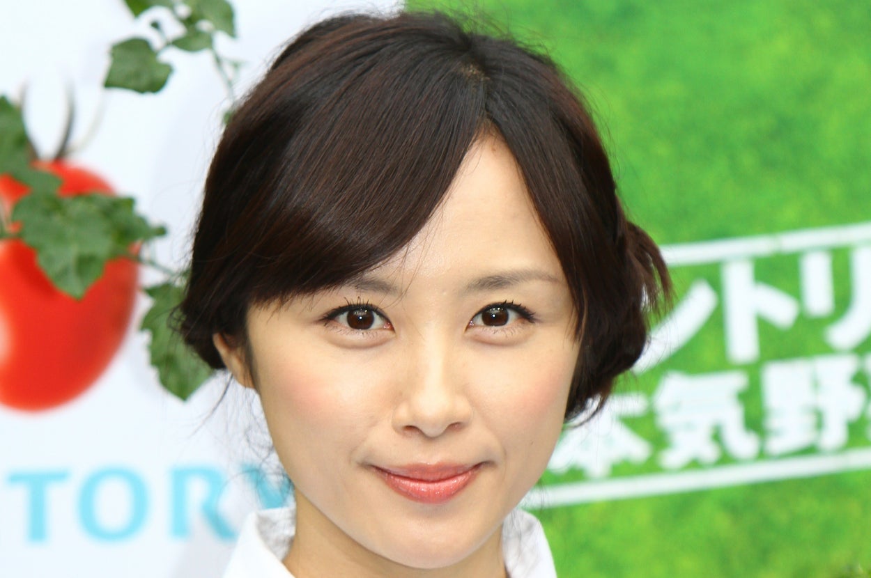 女性がカメラに向かって微笑んでいる。髪はアップにしており、白いシャツを着ている。背景には植物が見える。