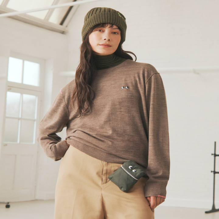 Person wearing a cozy sweater, beanie, and trousers, holding a small pouch with playful details, in a relaxed indoor setting