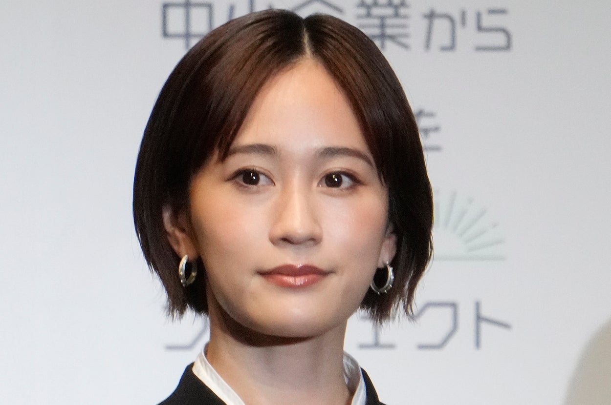 女性がフォーマルな服装でカメラを見つめる。背景に文字があるが詳細は不明。
