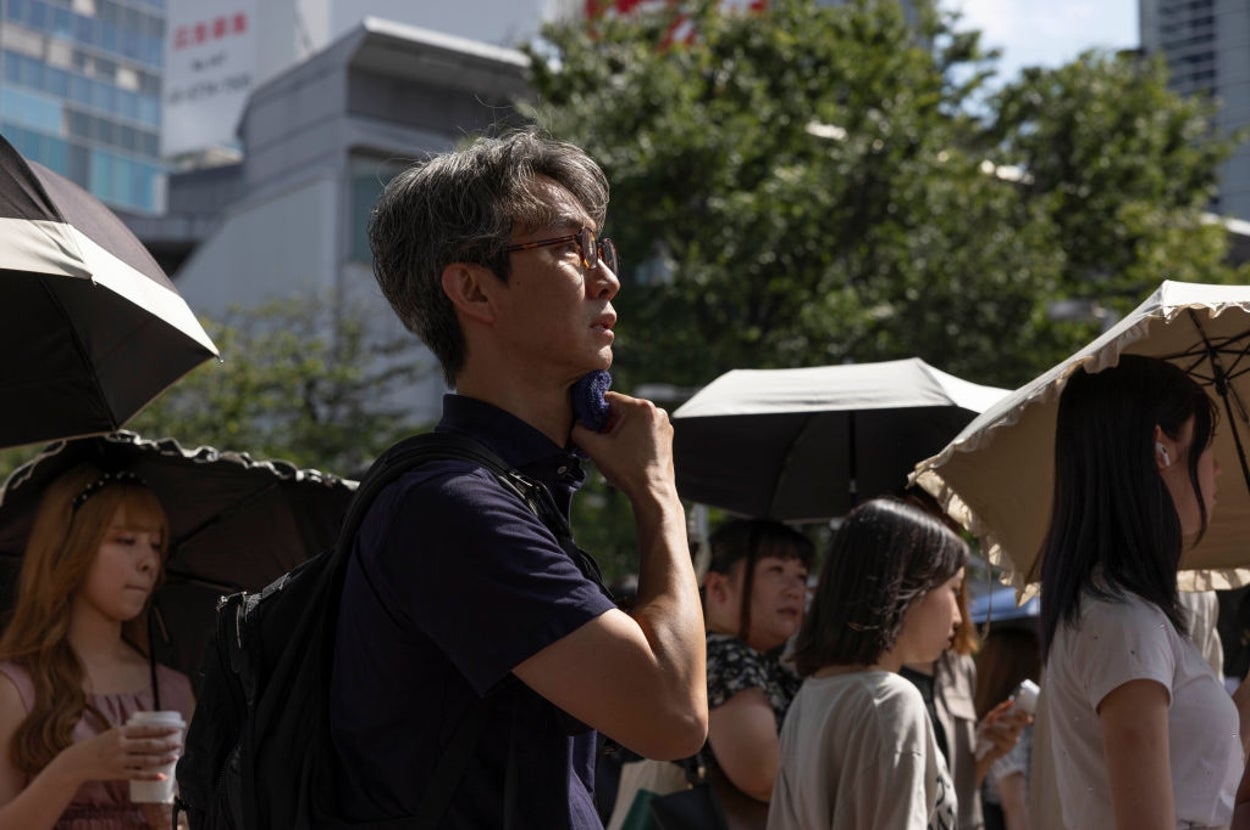 通りで日傘をさす人々の中、一人の男性がタオルで首を拭いている。