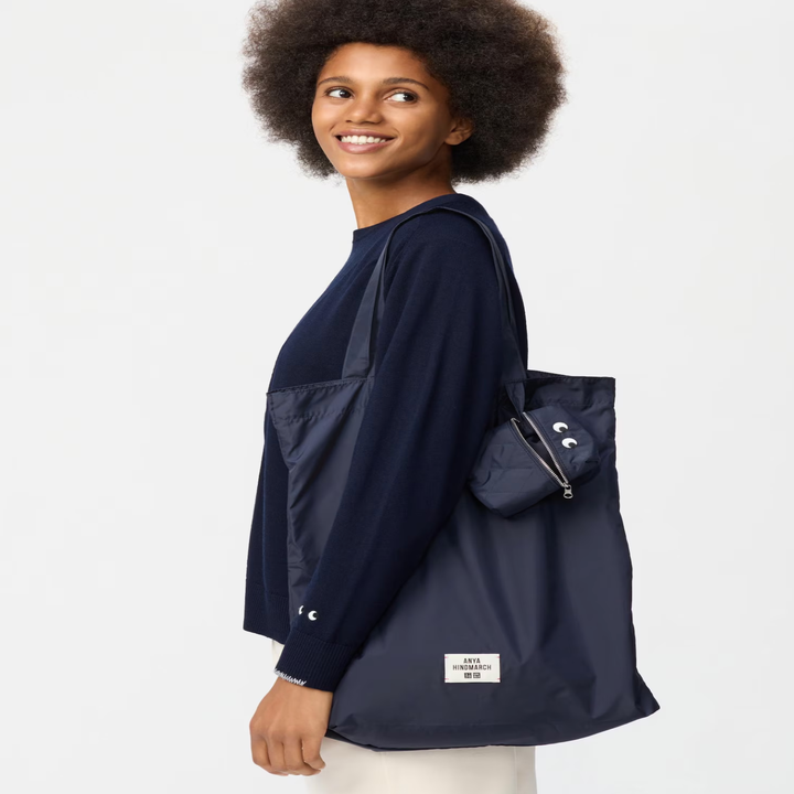A smiling person carries a sleek fabric tote bag with a pouch. The bag has a small label at the bottom