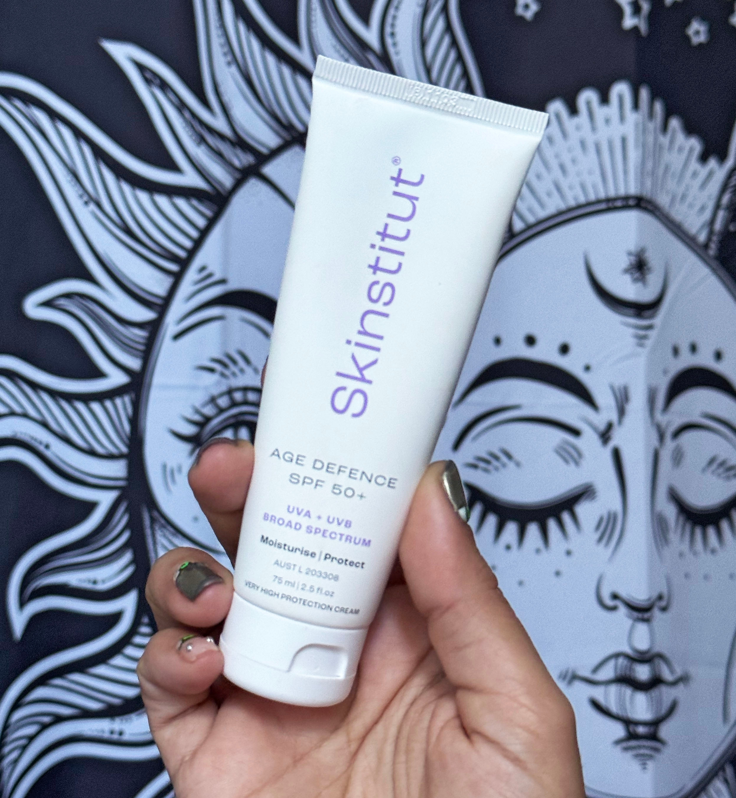 Person holding bottle of Skinstitut Age Defence SPF 50+ against a backdrop with celestial design