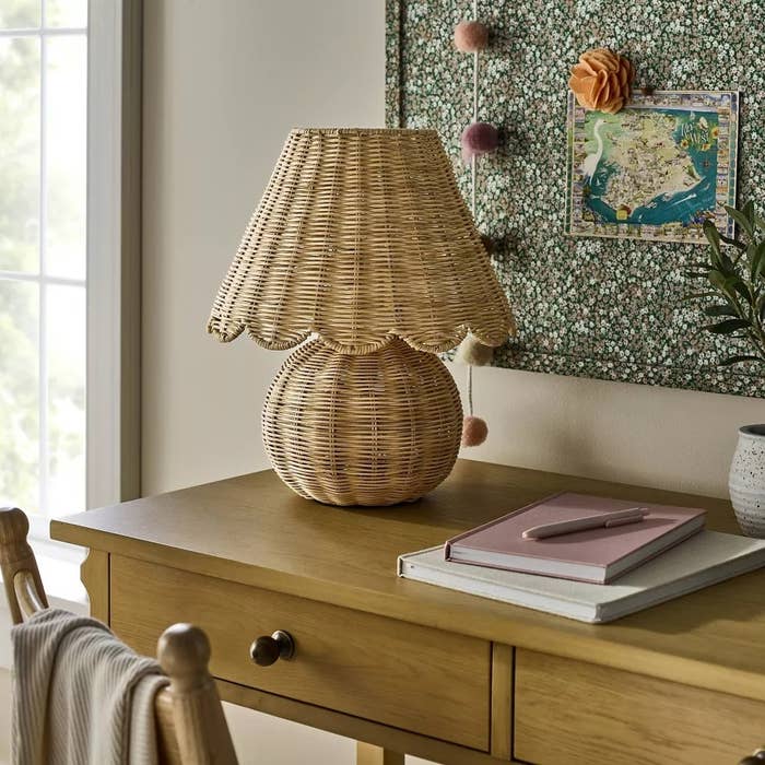 Woven rattan lamp with scalloped shadiness connected a woody desk, adjacent to a pinkish notebook. Corkboard with decorations successful the background