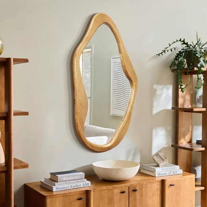 Decorative irregular-shaped reflector connected a wall, supra a woody sideboard with books and a bowl, flanked by shelves with plants and decor items