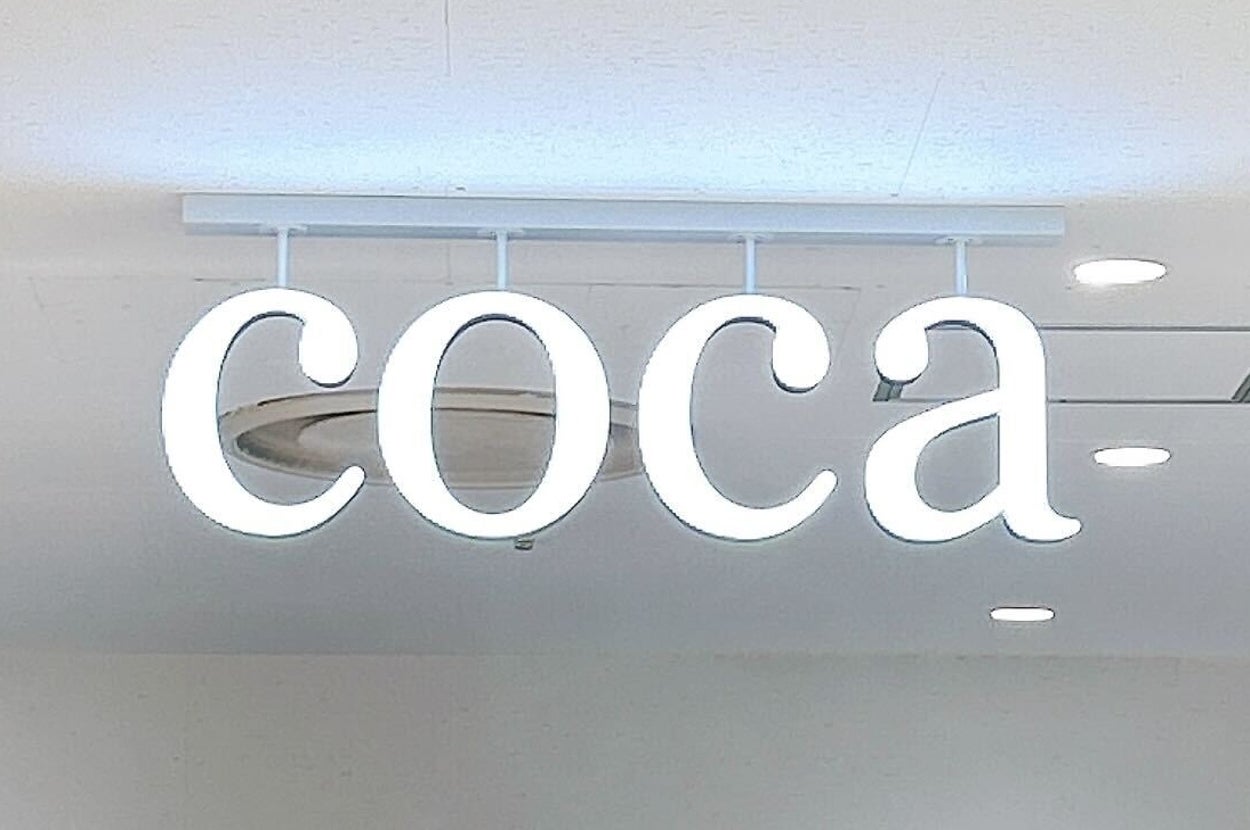 店内の天井から吊り下げられた「coca」の発光ロゴ看板。背景には洋服が並ぶラックが見える。