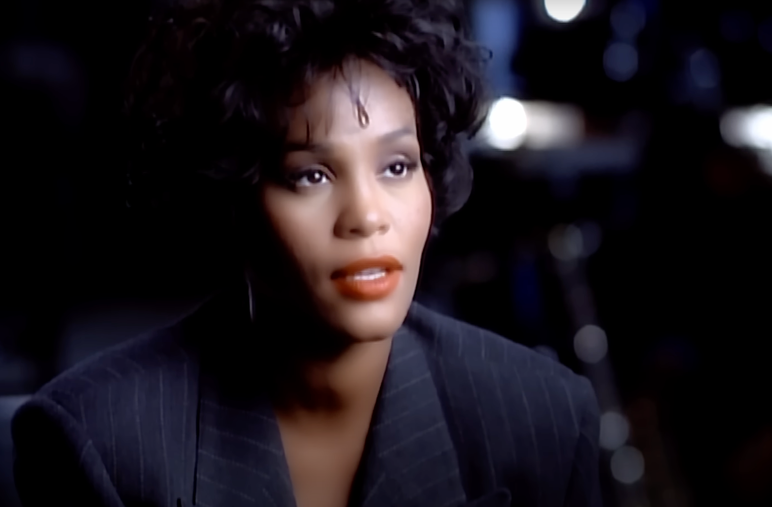 A person with curly hair wearing a pinstripe suit jacket, appearing to be in a TV or movie setting