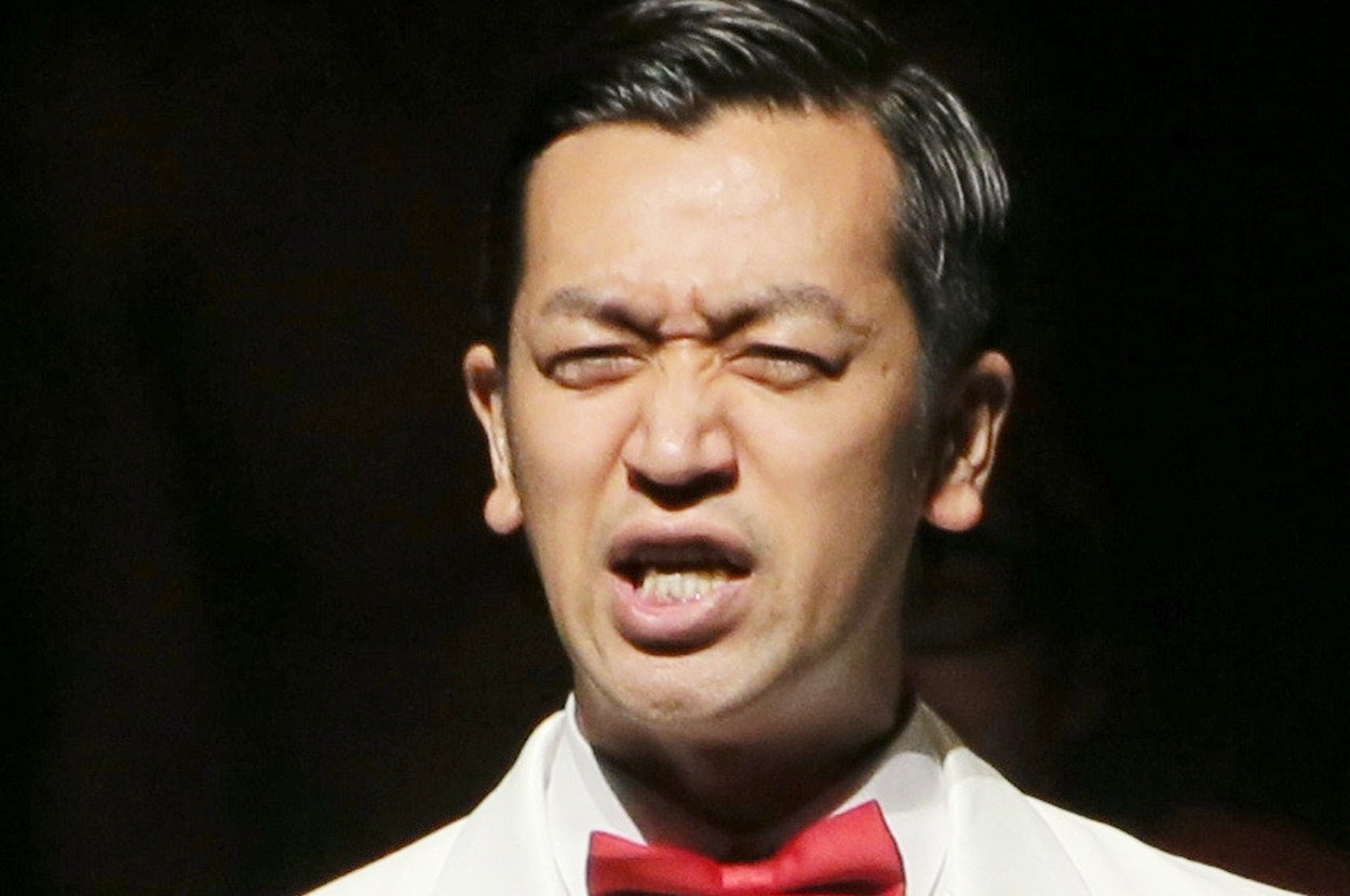 白いスーツと赤い蝶ネクタイを着た男性がステージでマイクを持って話す。背景には座っている人々がいる。