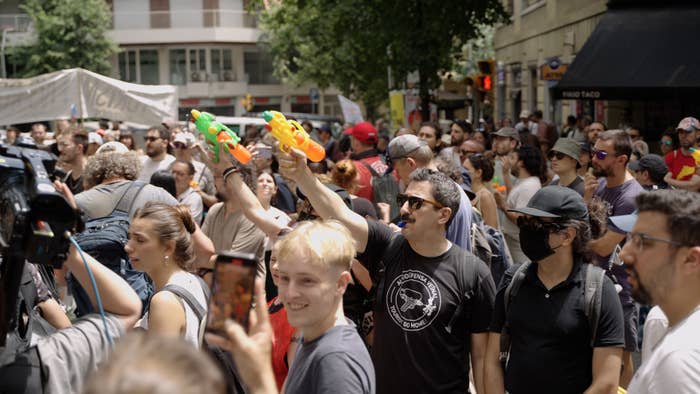 Crowd at a protest, some holding water guns. People are engaging peacefully and taking photos amidst the gathering