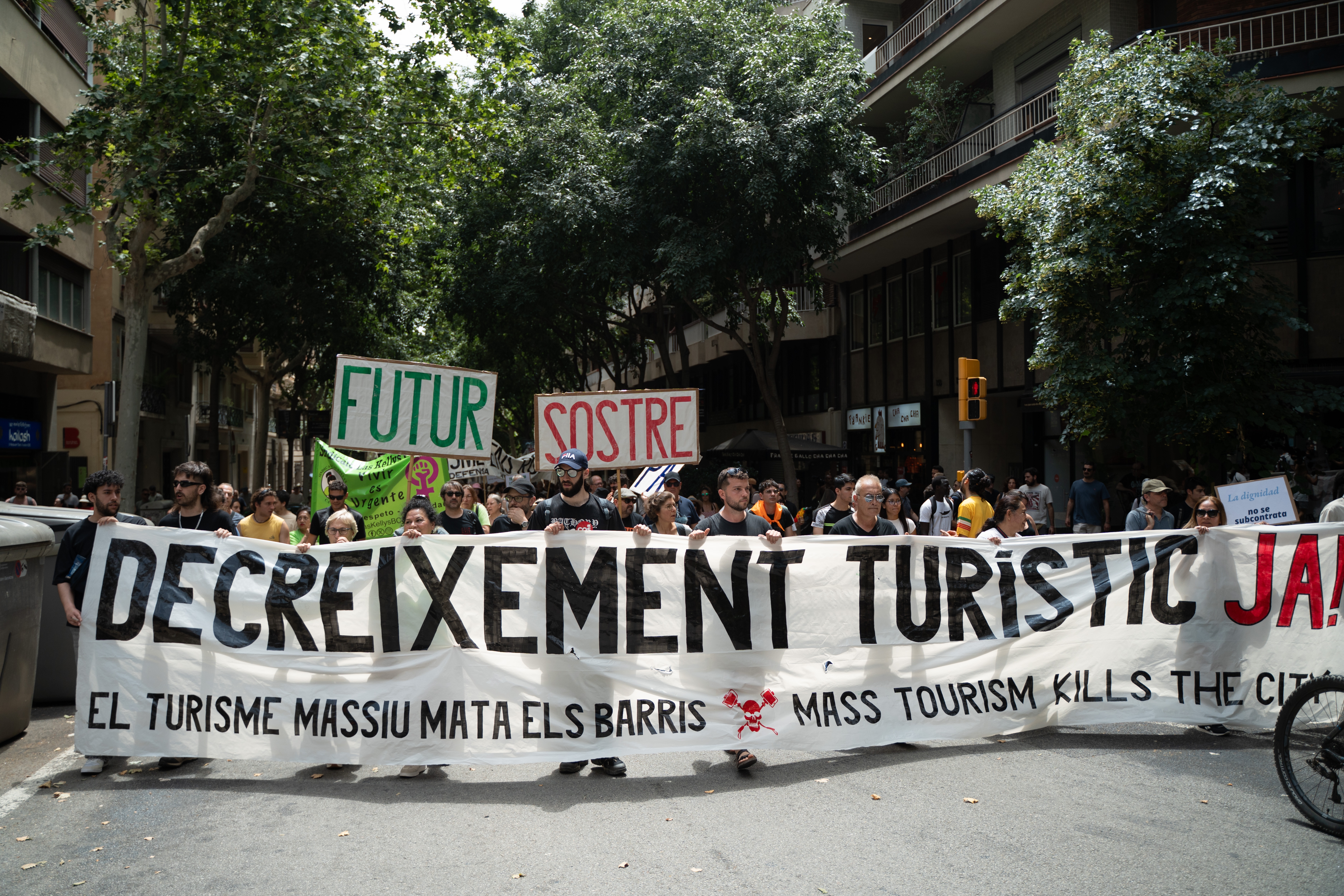 Protesters march holding banners against mass tourism, stating "Mass Tourism Kills the City" in a city street lined with trees