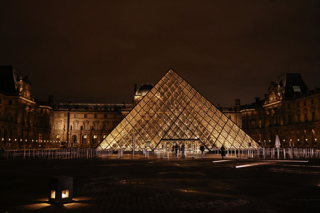 The Louvre Pyramid at night, illuminated and surrounded by the museum's historic buildings, creating a striking contrast