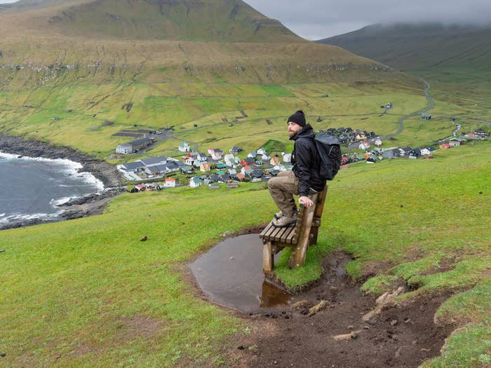 Person sits connected a woody seat overlooking a scenic coastal colony and rolling greenish hills. They deterioration casual, outdoor clothing