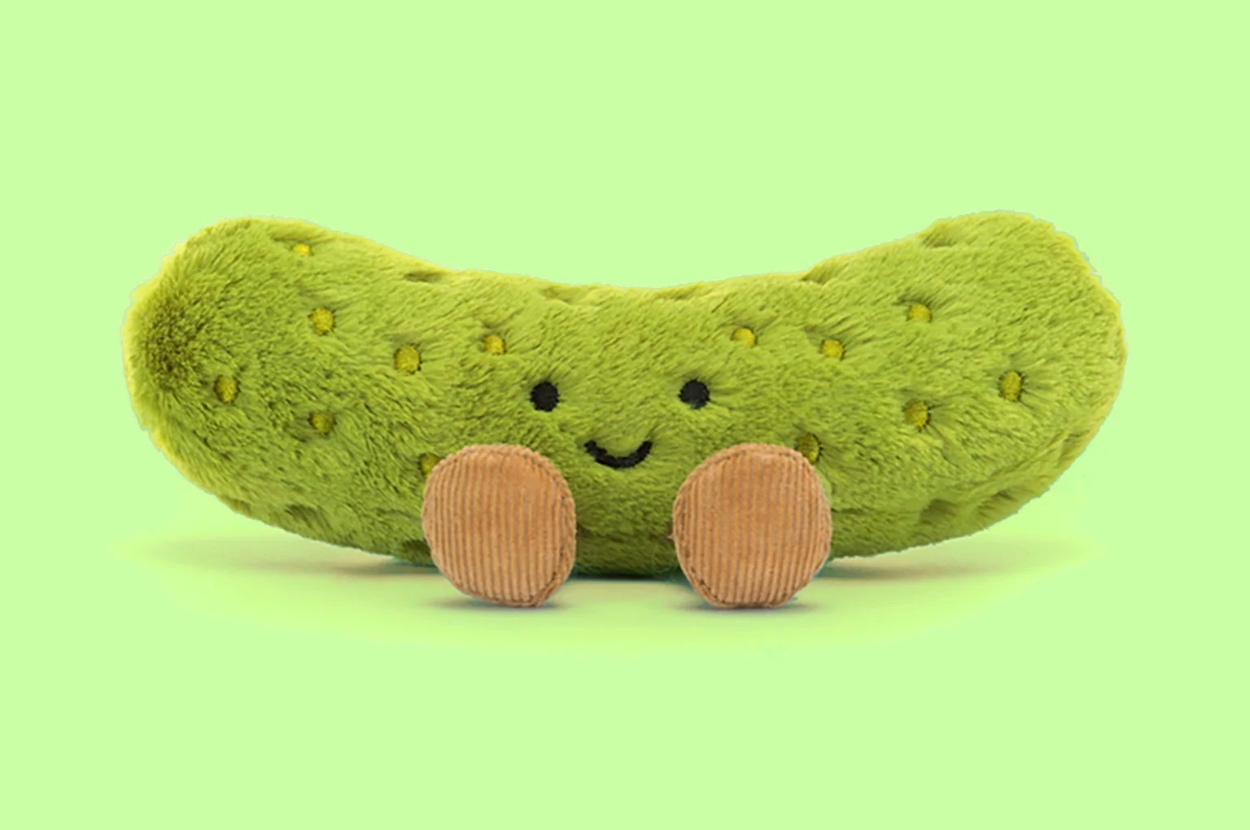 Plush pickle with a smiling face and two round feet, placed against a plain background