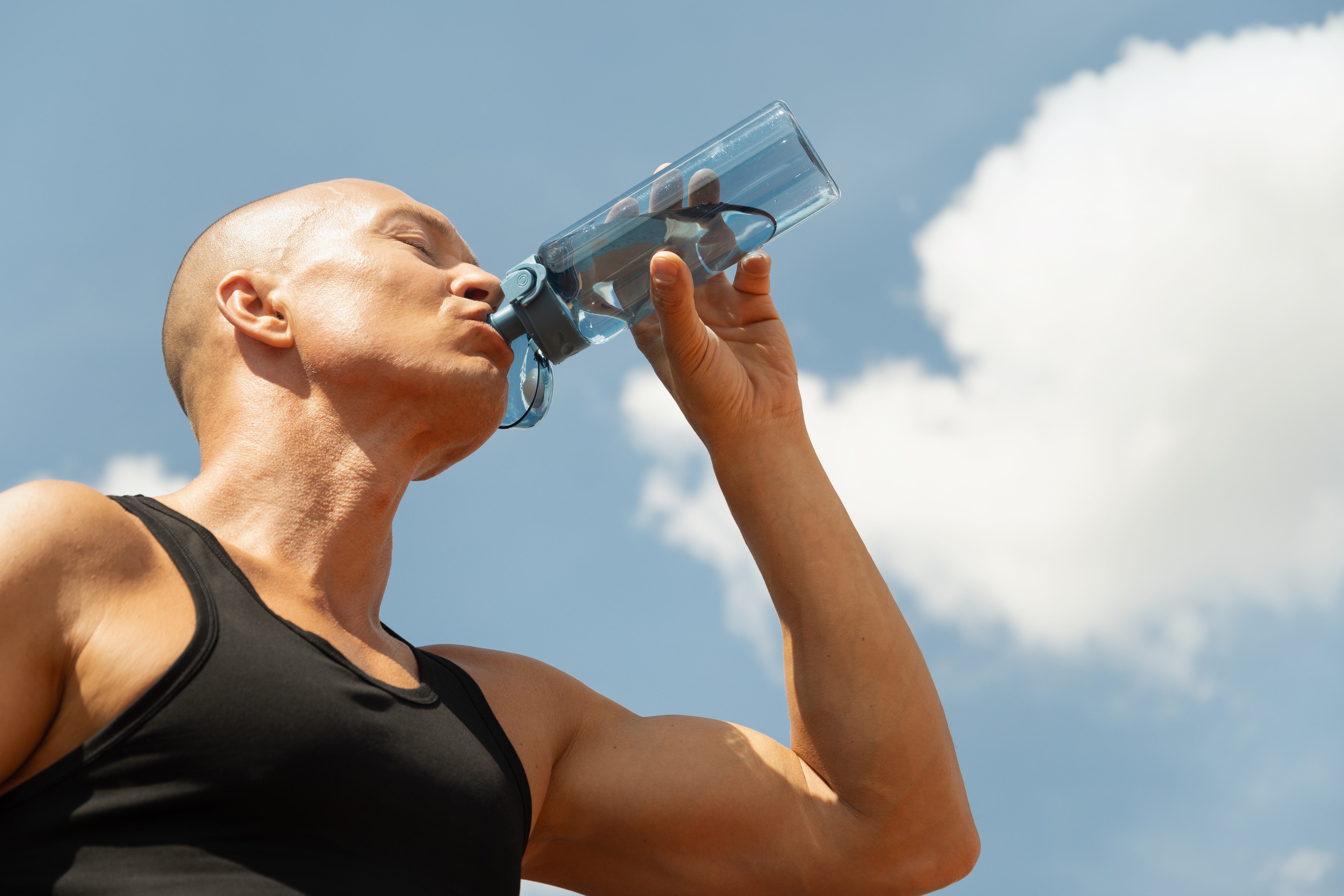 Person i en tank top som drikker fra en vannflaske under en klar himmel