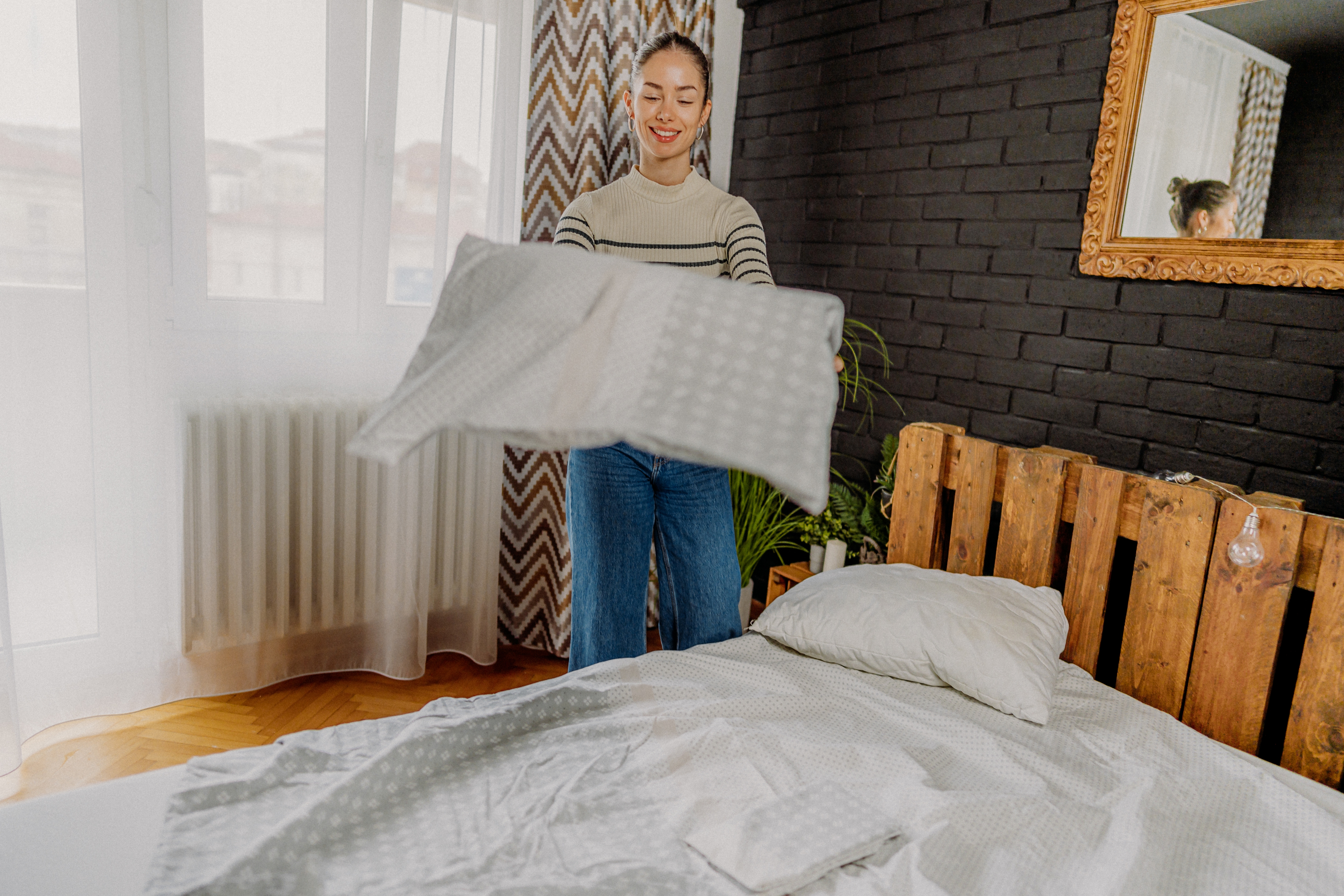 Person i en stripet genser og jeans som lager en seng i et koselig rom med en pallhodegavl og et speil på en svart murvegg