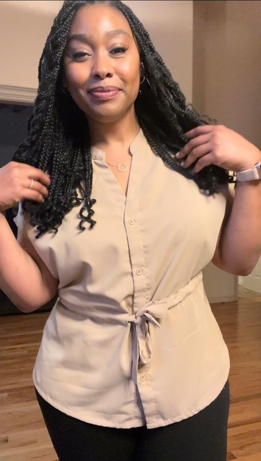 Person wearing a belted beige blouse and black pants poses confidently indoors, smiling and adjusting hair with hands