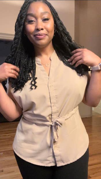 Person wearing a belted beige blouse and black pants poses confidently indoors, smiling and adjusting hair with hands