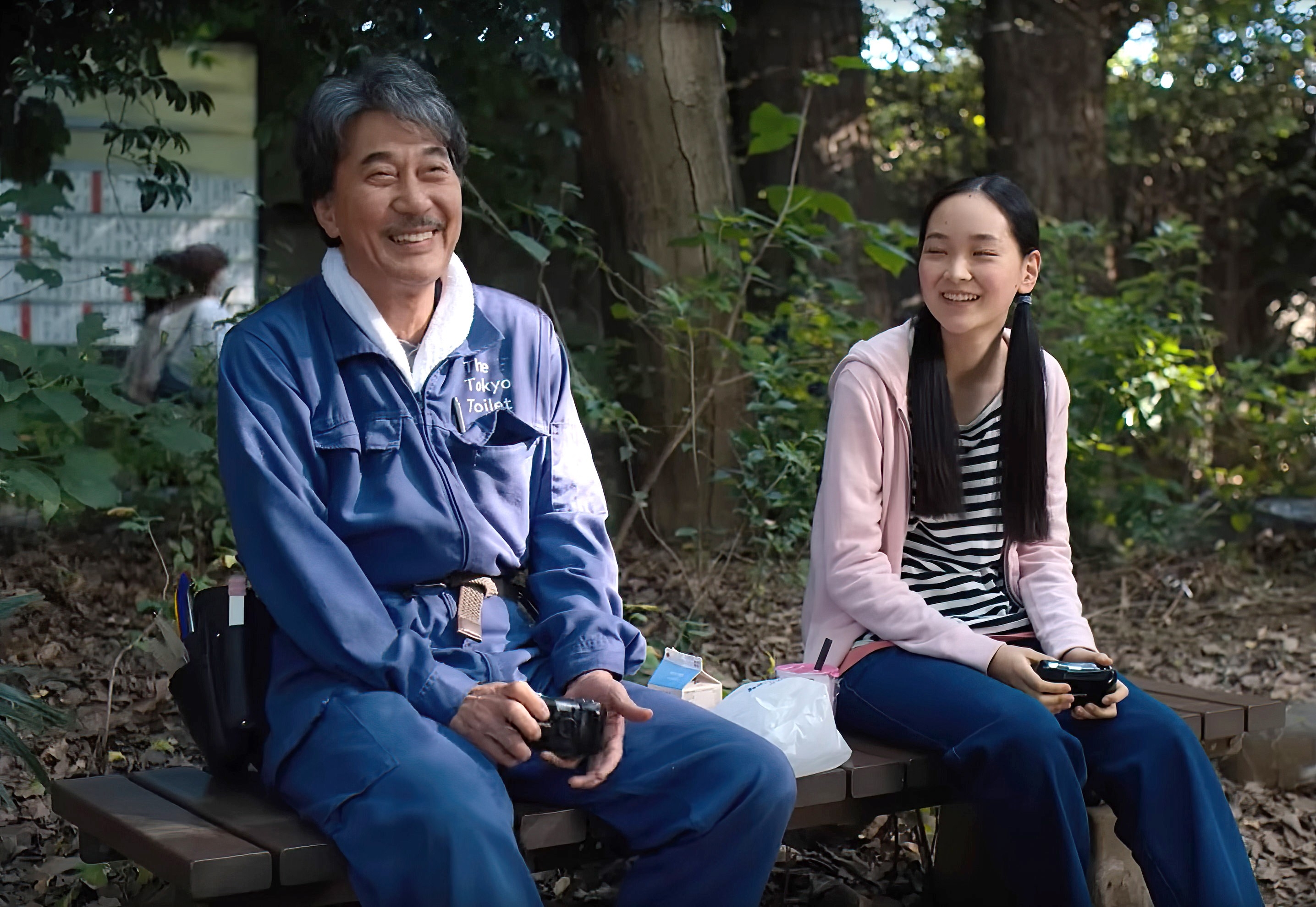 Two radical   sitting connected  a seat  outdoors, smiling and holding gaming controllers, with trees successful  the background