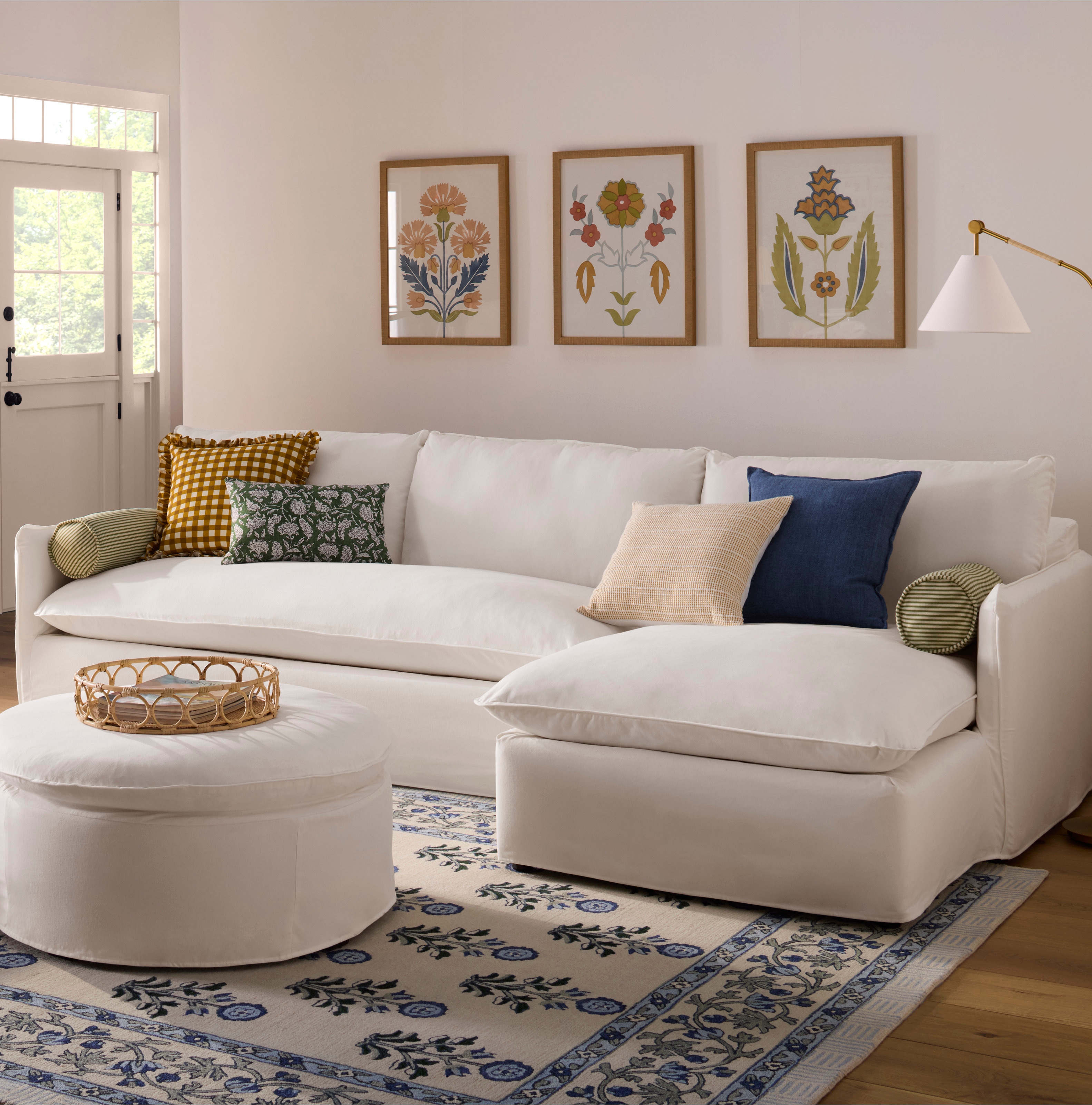 A cozy living room features a white sectional sofa with assorted pillows, a round ottoman, and botanical wall art, ideal for home decor shopping