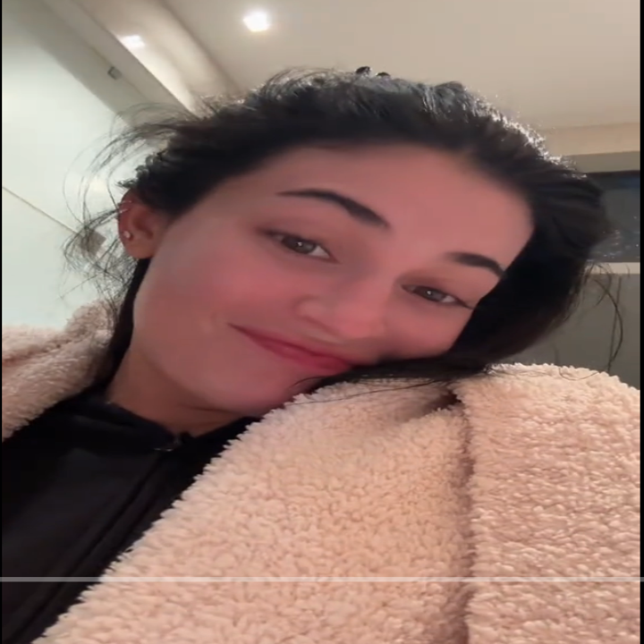 Person taking a selfie in a cozy, textured jacket indoors, smiling softly at the camera