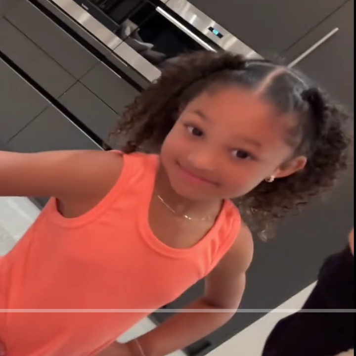 Young child with curly hair and an orange sleeveless top smiles confidently indoors