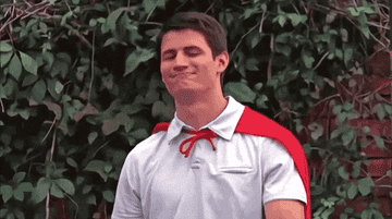 Person nodding and smiling confidently, wearing a casual shirt with a red cape tied around their neck, standing in front of leafy vines