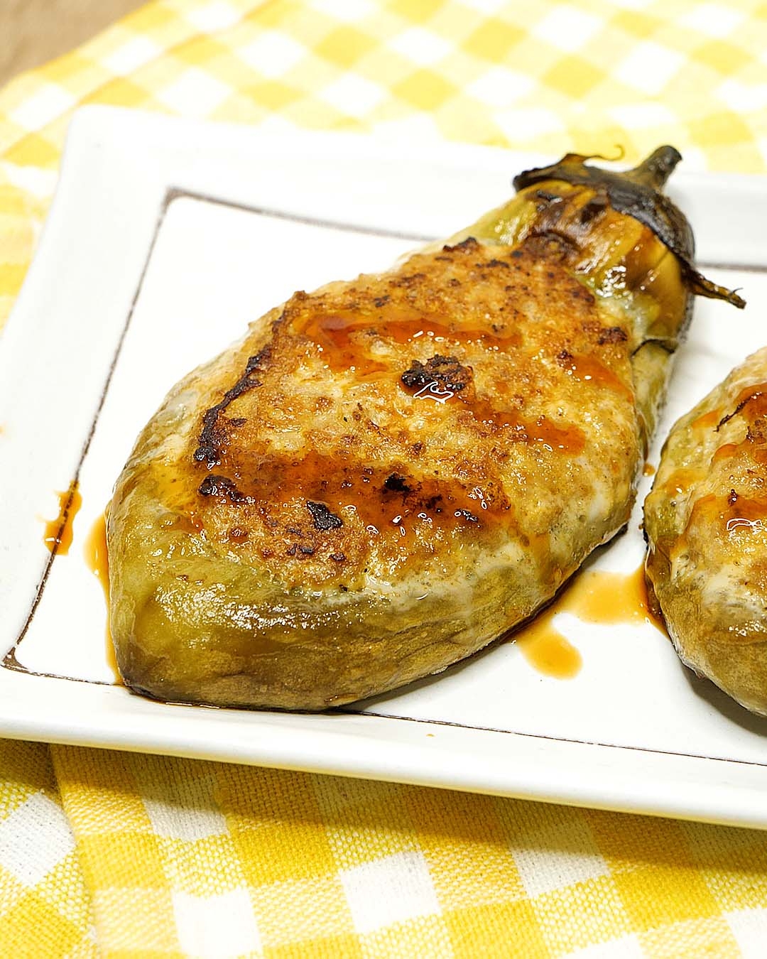 焼かれたナスが白い皿にのっている。ナスにはソースがかかっていて、背景に黄色のチェック柄の布が見える。