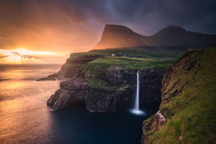 Dramatic scenery of a waterfall flowing into the ocean, with steep cliffs and a mounting prima illuminating a mountainous backdrop