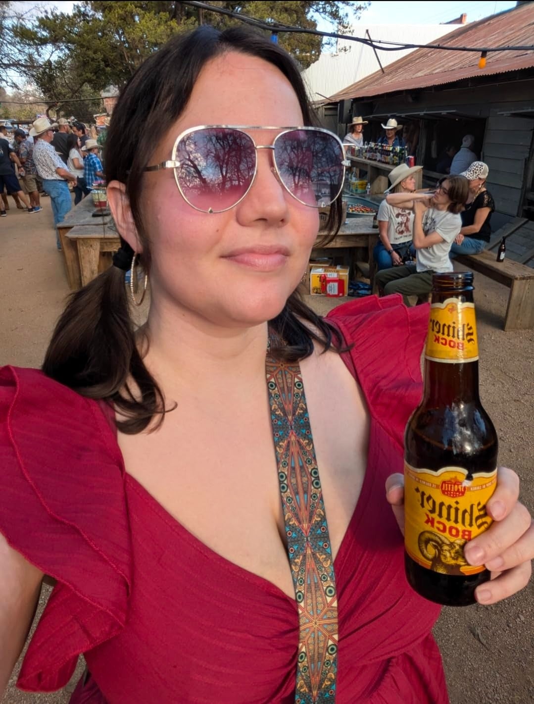 Person outdoors holding a Shiner Bock beer, wearing sunglasses and a vibrant, ruffled formal  with puffy sleeves. People are enjoying a casual gathering