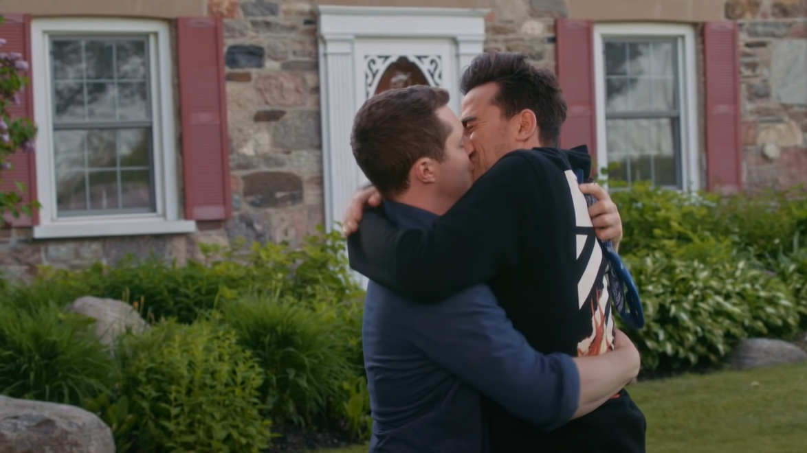 Patrick and David kiss passionately, arms around each other, in front of a stone house