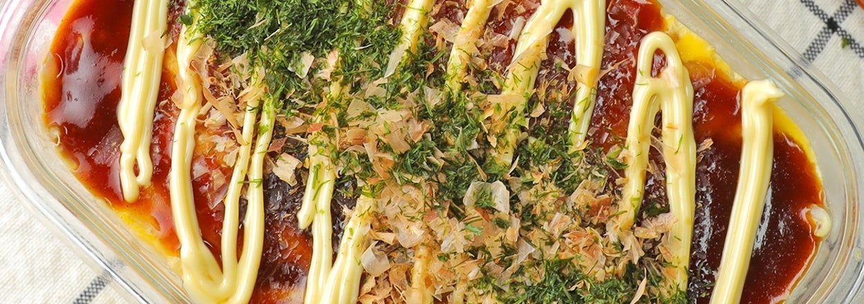 ガラス容器に入ったお好み焼き風チーズ焼き。マヨネーズ、ソース、青のり、かつお節がかけられている。横に木製スプーン。