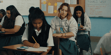 Students in a classroom taking a test, with one student, wearing a hoodie, glancing at another&#x27;s paper