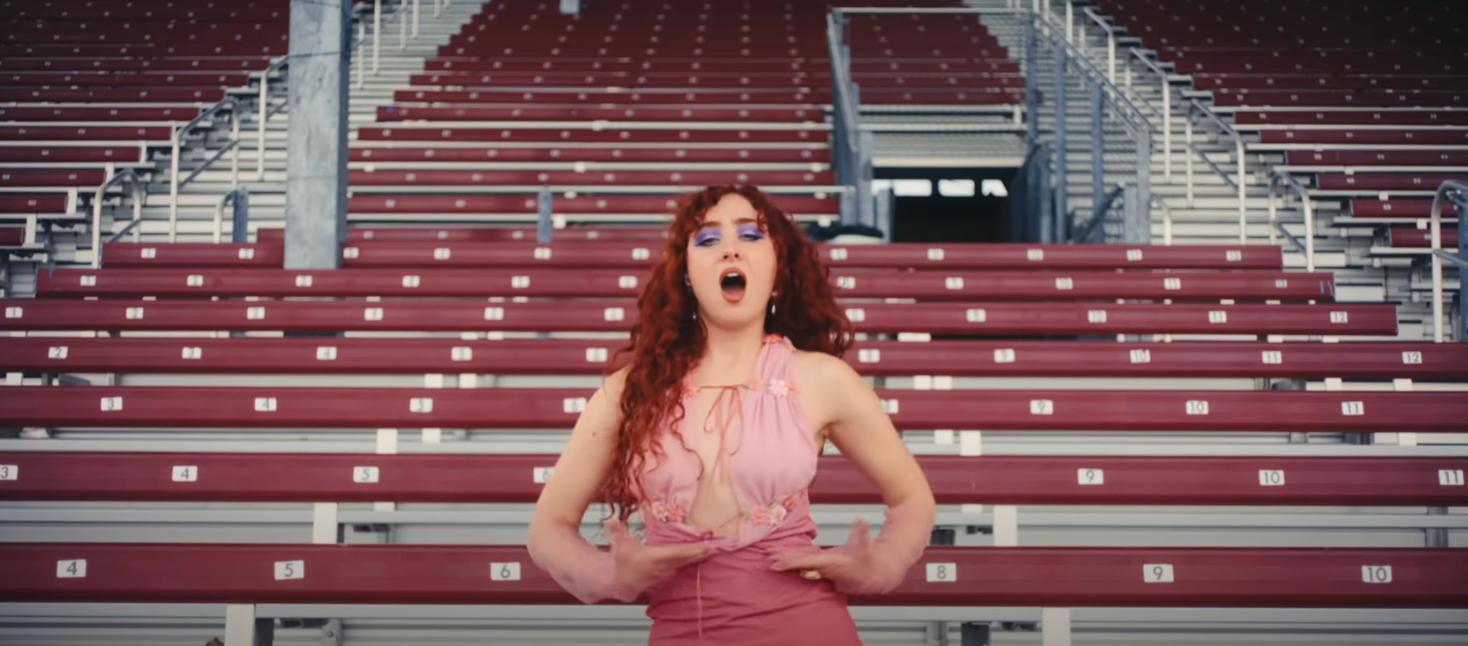 A person with curly hair sings passionately in an empty stadium, wearing stylish clothing with a lace-up design
