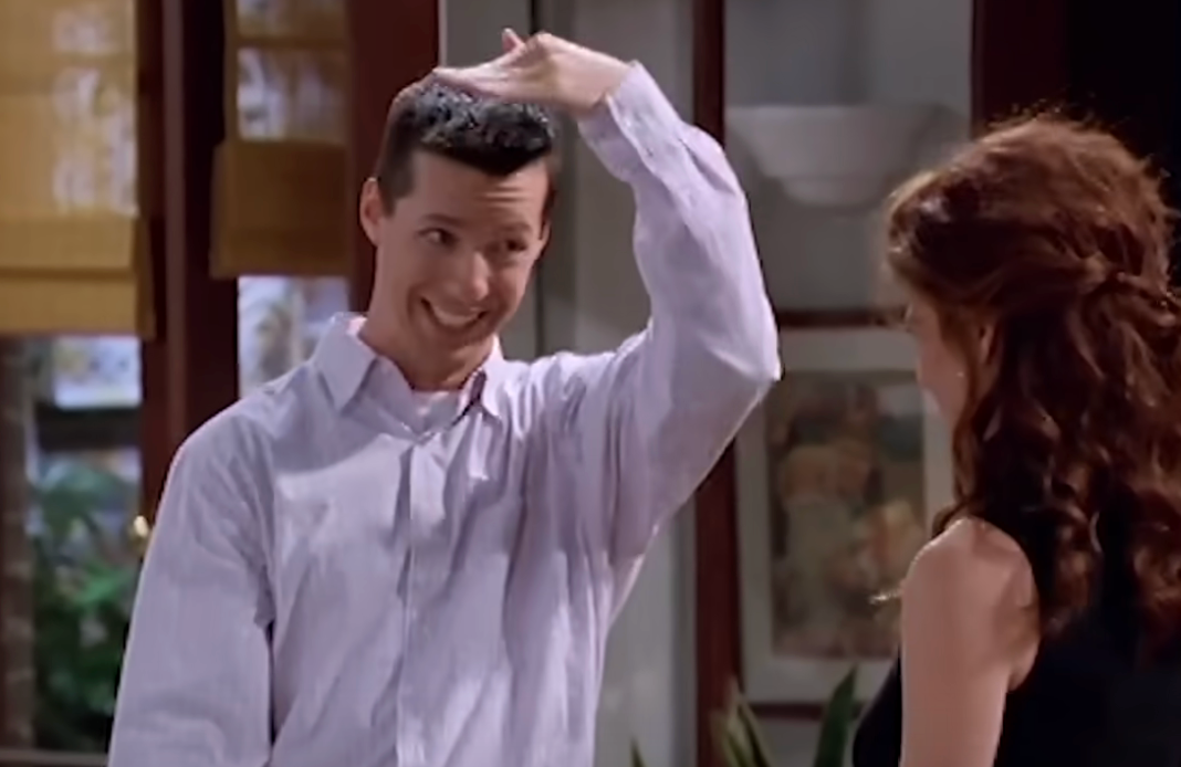 A character with a playful expression gestures to their hair while speaking with another person in a sitcom scene