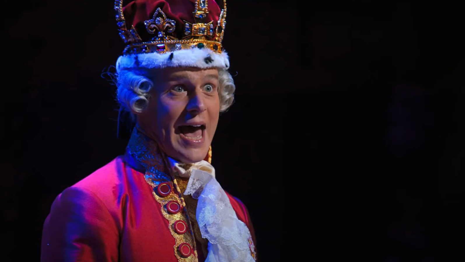Person in elaborate period costume, including a crown, performing on stage with expressive facial expression