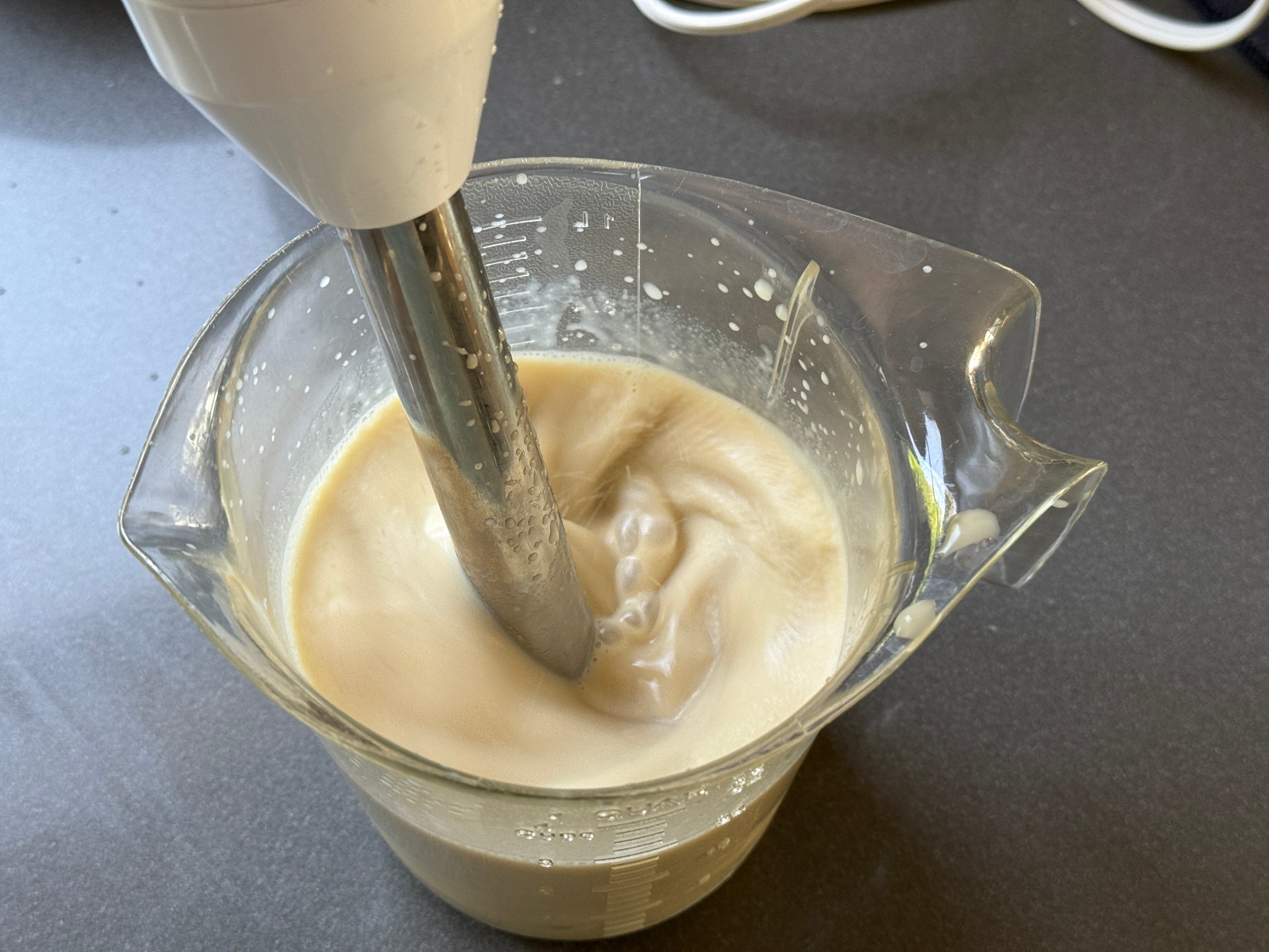 A handheld blender mixing creamy batter in a clear measuring jug on a countertop