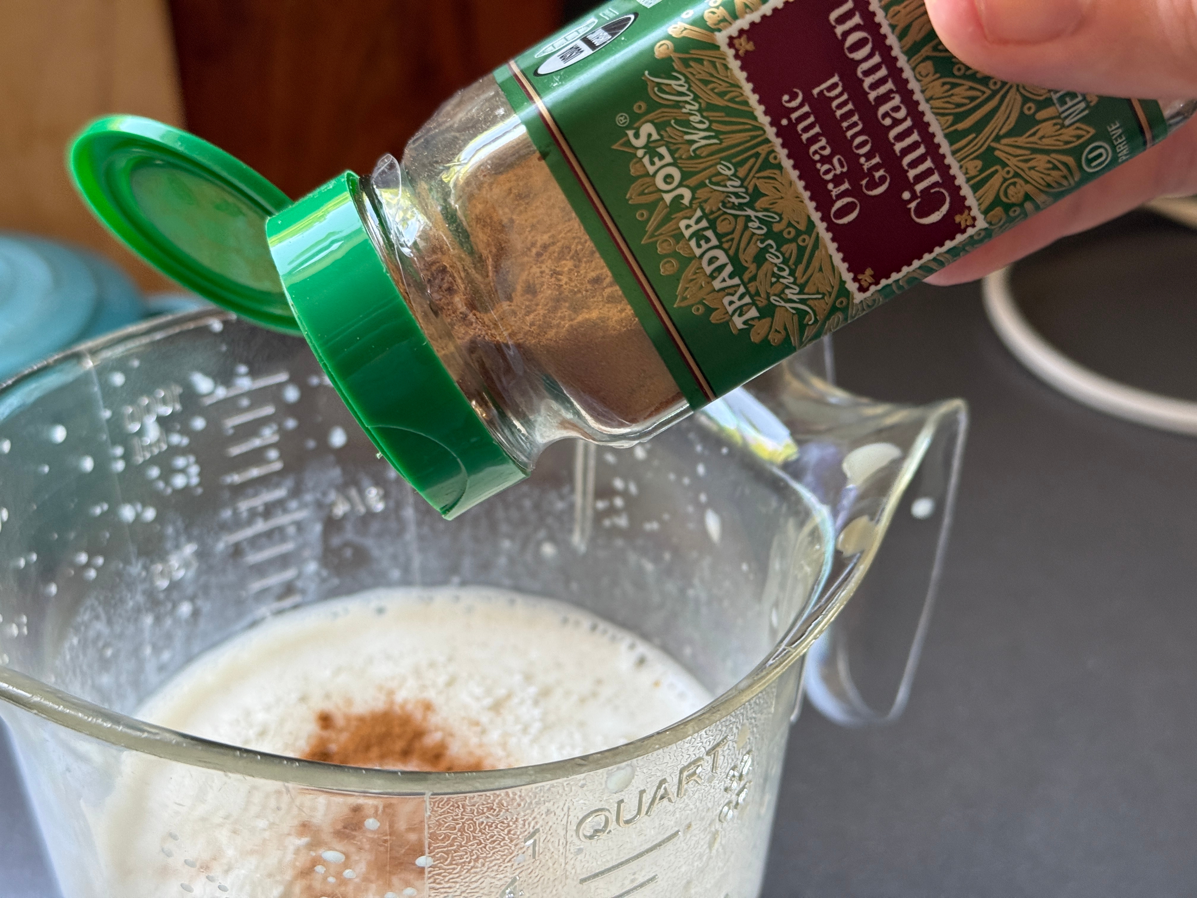Cinnamon is being sprinkled into a glass measuring cup containing a frothy liquid