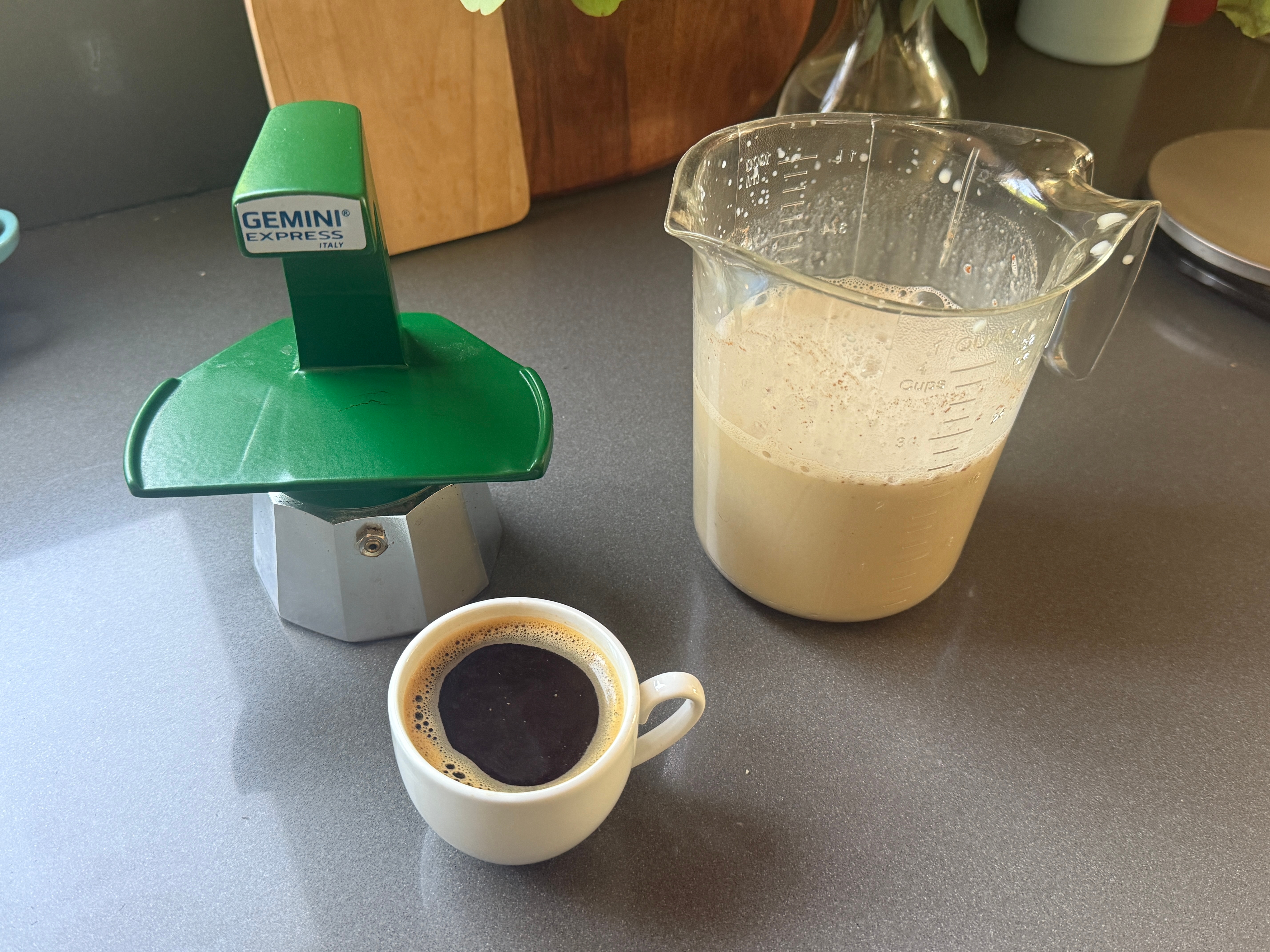 Espresso maker, cup of espresso, and a measuring pitcher with frothy liquid on a kitchen counter